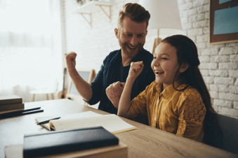 A smiling father and daughter at a desk with fists raised in celebration over an open notebook.