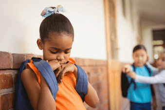 A young girl in an orange top and backpack leans against a brick wall looking down, while two children talk in the blurred background.