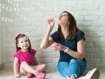 A Woman Sitting And Blowing Bubbles While A Little Girl Sits Beside Her Watching And Smiling