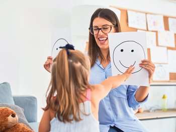 A Woman Smiling And Holding Up A Piece Of Paper With A Smiley Face And Another One With A Frowny Face While A Little Girl Points To The Paper With The Smiley Face