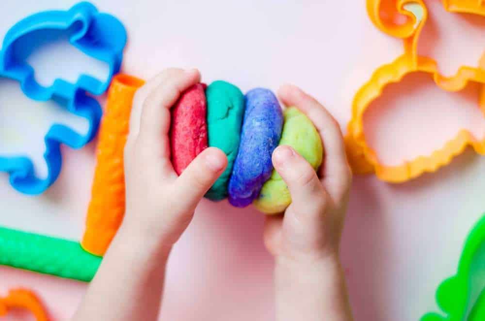 Toddler With Autism’s Hands Playing With Colorful Playdough With Toys In Background