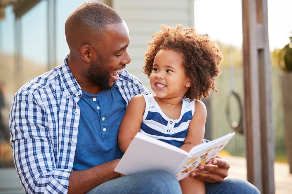 Father Laughing And Reading A Book To His Daughter