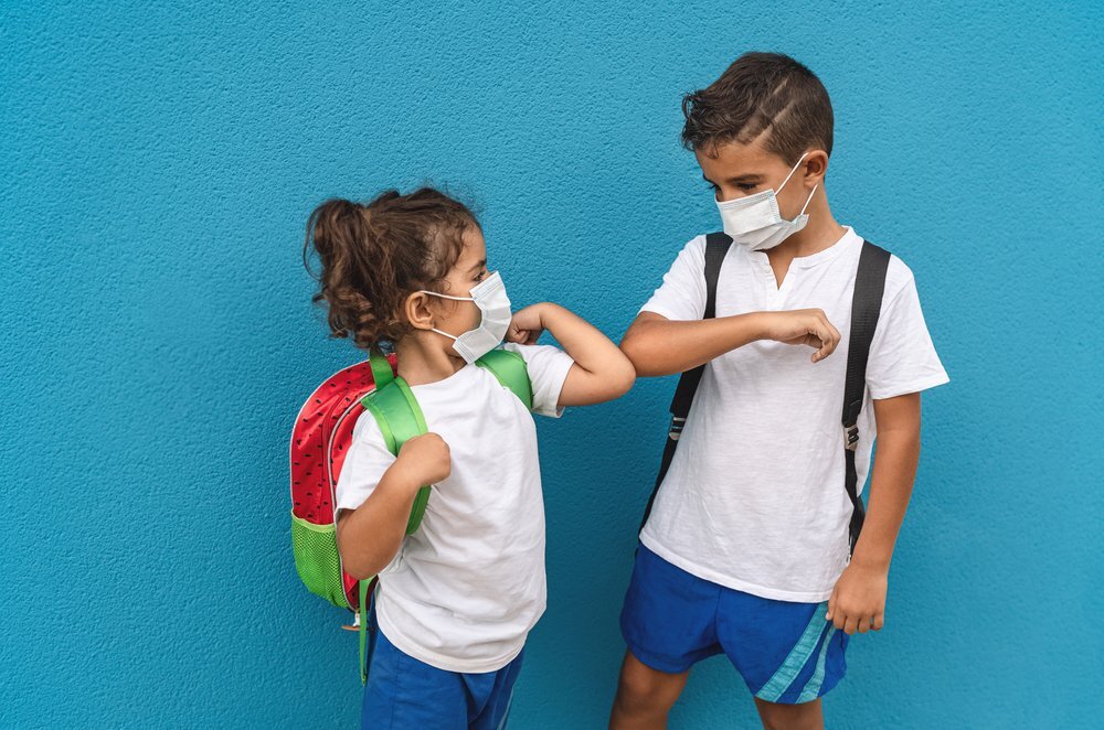 Two Kids In Masks Bumping Elbows In Front Of A Blue Wall