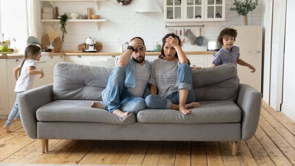 Mother And Father Overwhelmed With Parenting On A Couch With Children Running Around Them