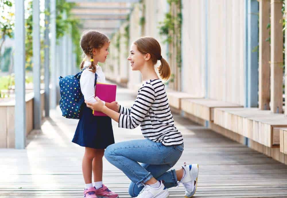 Parent Leaning Down To Talk To Child With Autism Wearing A Backpack Before Dropping Off At School