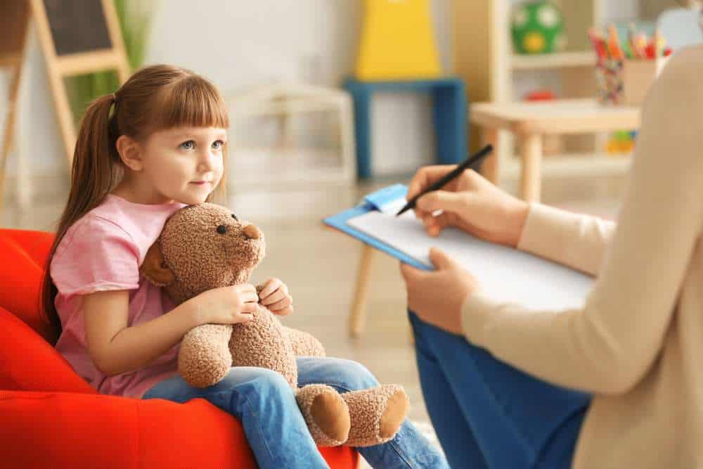 Young Girl Holding A Teddy Bear While Talking To A Psychologist Holding A Clipboard