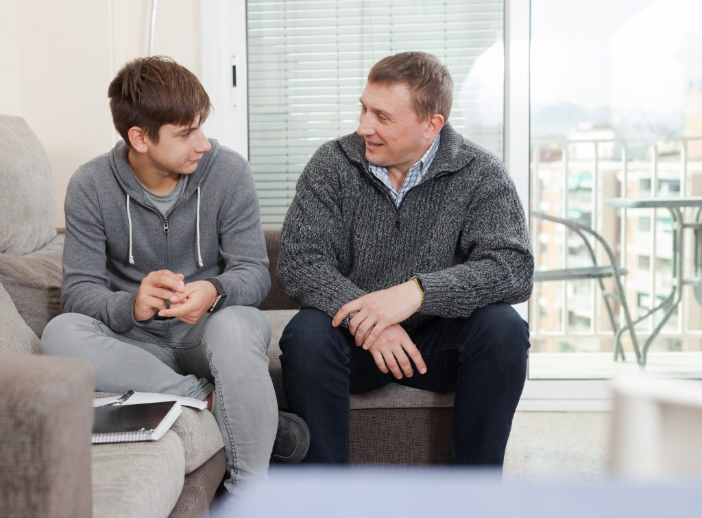 A Father Sitting On A Couch And Talking To Their Teenage Son