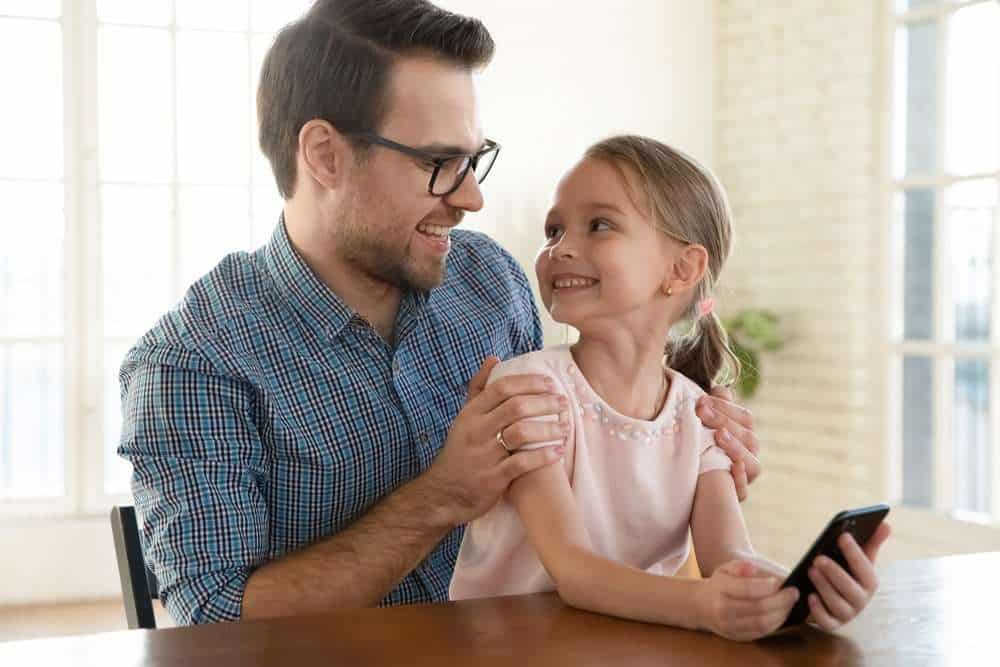 Father Hugging and giving noncontingent attention to his Daughter While She Smiles And Holds A Phone