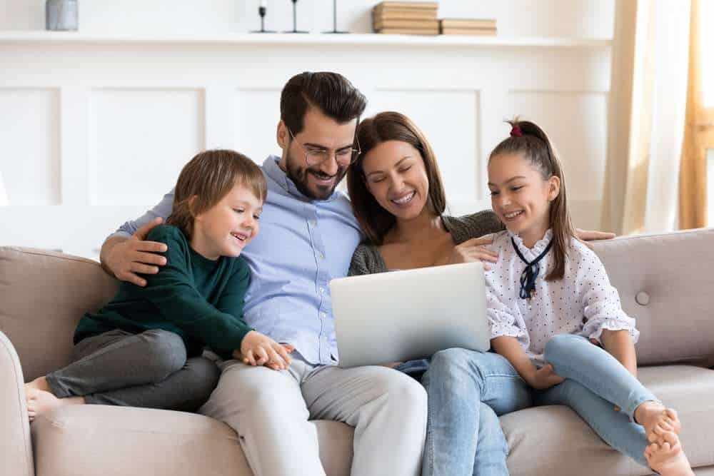 Mother, Father, Son, And Daughter Laughing At  Laptop On A Couch
