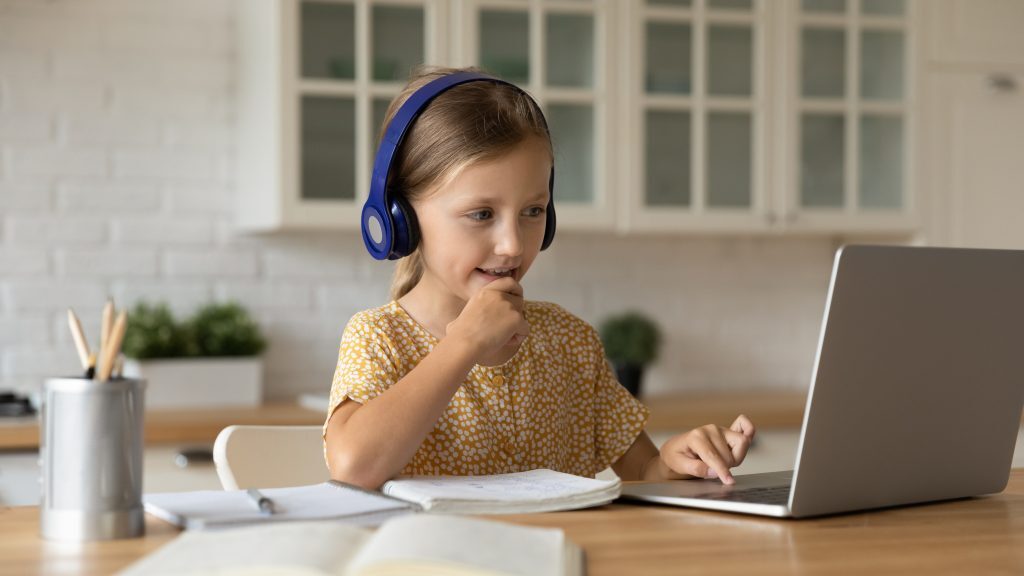 A Little Girl Sitting At A Desk Wearing Headphones And Looking At A Computer With A Notebook And Pens Next To Her