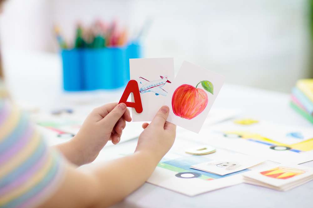Child Holding A Picture Of An Airplane, Apple, and the Letter A