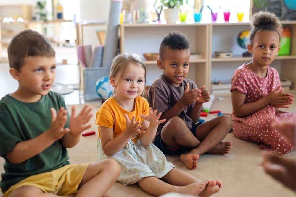 Four Children Sitting On The Ground And Clapping Their Hands During Kindergarten Class