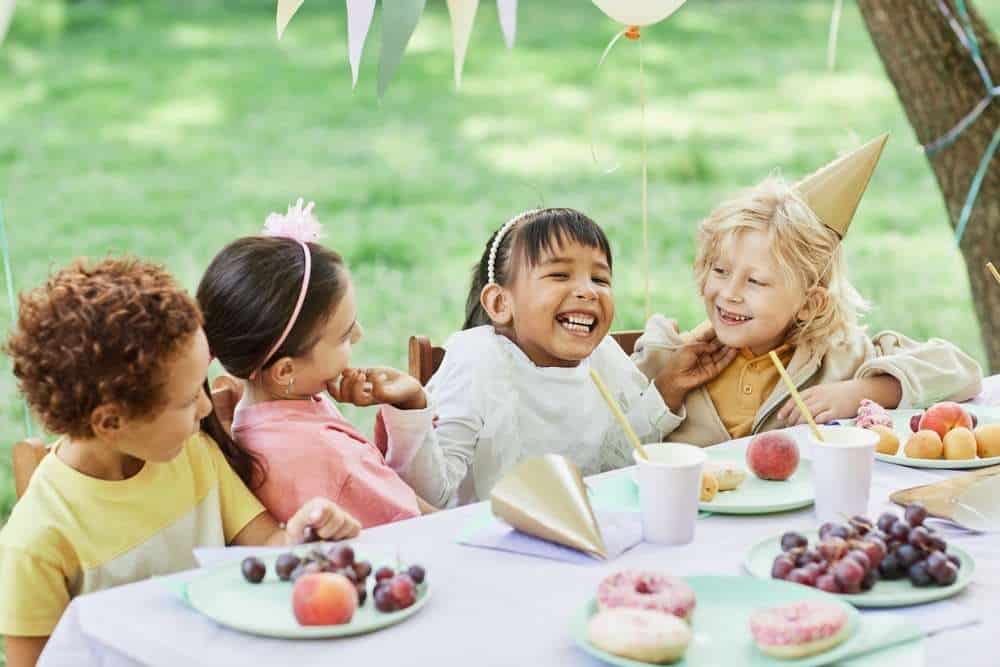 Four Kids At A Birthday Party Eating Snacks At A Table And Laughing