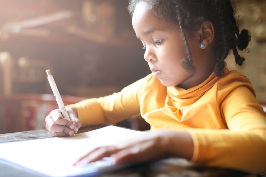 A Little Girl Sitting At A Desk And Writing In A Notebook