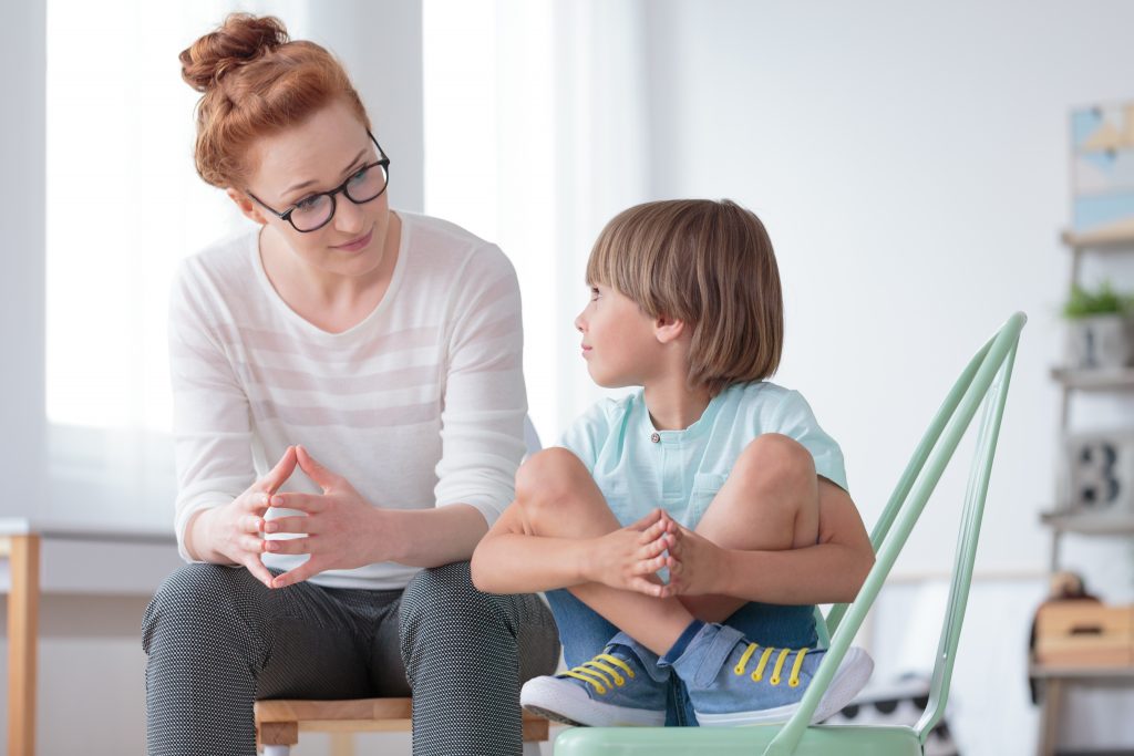 ABA Therapist Talking To A Child With Autism Who Is Sitting In A Chair And Hugging Their Knees